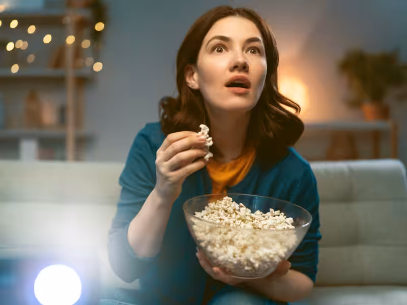 A woman with dark hair sits on a couch, holding a bowl of popcorn and looking intently ahead, appearing surprised or captivated, with cozy lights and home decor in the background.