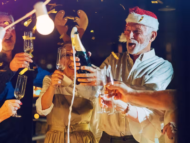 A group of people celebrate together, raising champagne glasses. One man wears a Santa hat and pops a bottle, while a woman beside him wears reindeer antlers. Confetti falls around them in a festive, lively scene.