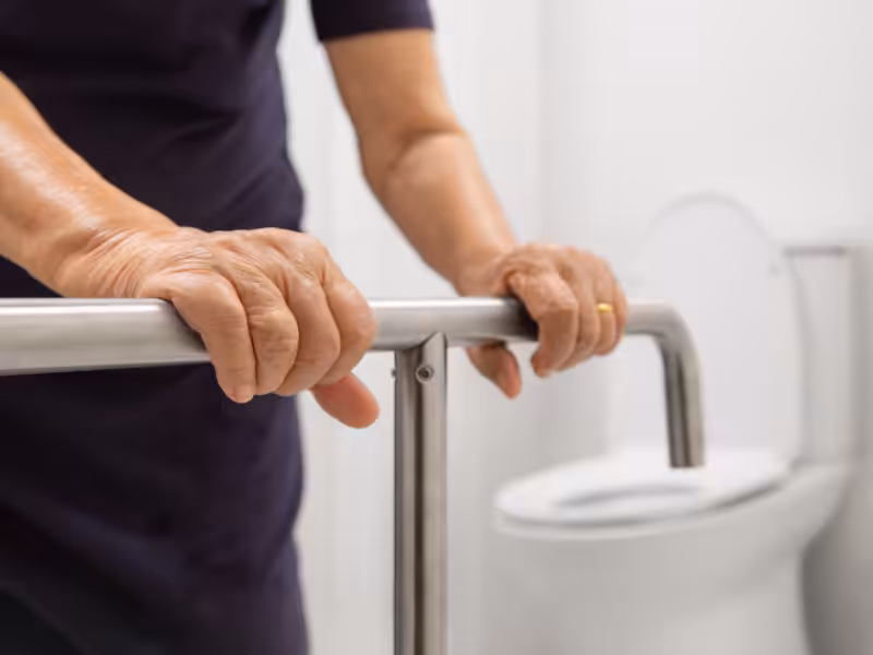 An elderly person gripping a support rail next to a toilet in a bathroom, suggesting assistance with mobility or safety while using the restroom.