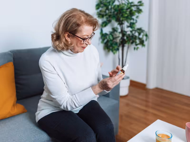 An older woman with glasses sits on a sofa, looking closely at a medication bottle she is holding. There is a plant behind her and a small table with a glass in front of her.