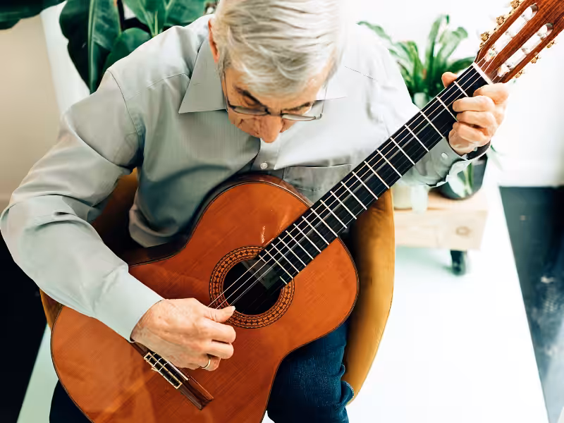 An elderly man with gray hair and glasses is sitting and playing a classical guitar, surrounded by green houseplants.