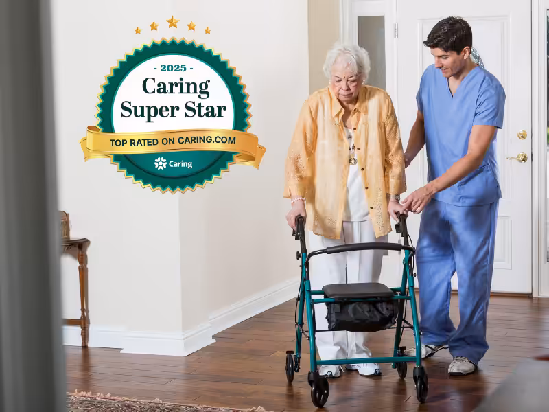 A young male caregiver in blue scrubs assists an elderly woman using a walker indoors. A 2025 Caring Super Star award badge from Caring.com is displayed on the left side of the image.