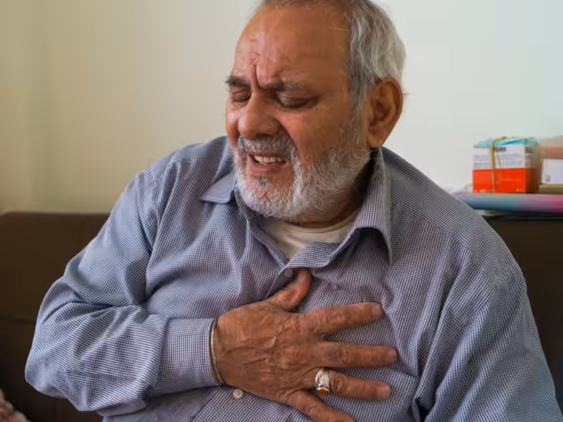 An elderly man with gray hair and a beard sits on a couch, clutching his chest with a pained expression, possibly experiencing chest pain or discomfort.