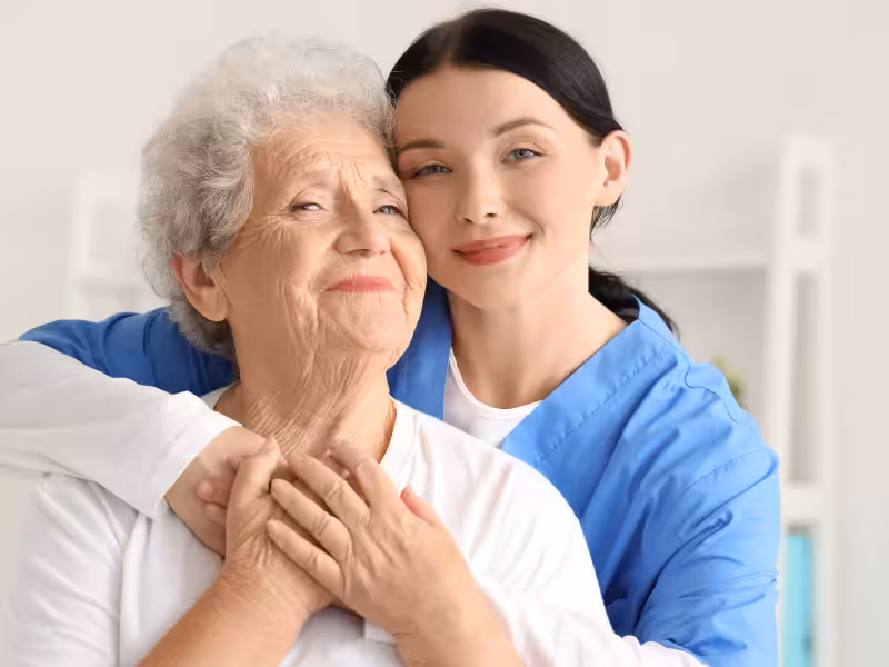 A smiling elderly woman is embraced from behind by a young female caregiver in blue scrubs. They look content and happy, standing together in a bright room.