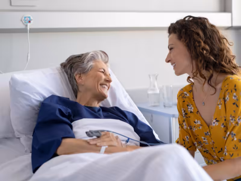 An older woman lies in a hospital bed smiling at a younger woman in a yellow dress, who sits beside her holding her hand. The scene appears warm and supportive.