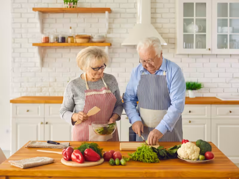 An elderly couple wearing aprons prepares a meal together in a bright kitchen. The man chops vegetables while the woman holds a bowl and a spatula. Fresh produce, including peppers and lettuce, sits on the wooden counter.