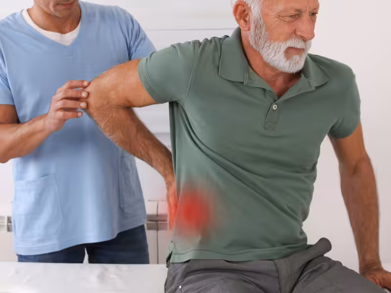 An older man in a green shirt sits on an exam table, holding his lower back in pain while a healthcare professional in blue scrubs examines him. A red highlight marks the area of discomfort on his lower back.