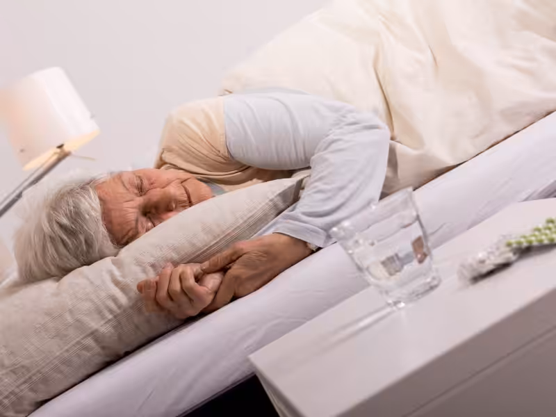 An elderly woman with gray hair sleeps on her side in bed, covered with a cream blanket. On the bedside table are a glass of water and some medication. A lamp is also visible in the background.