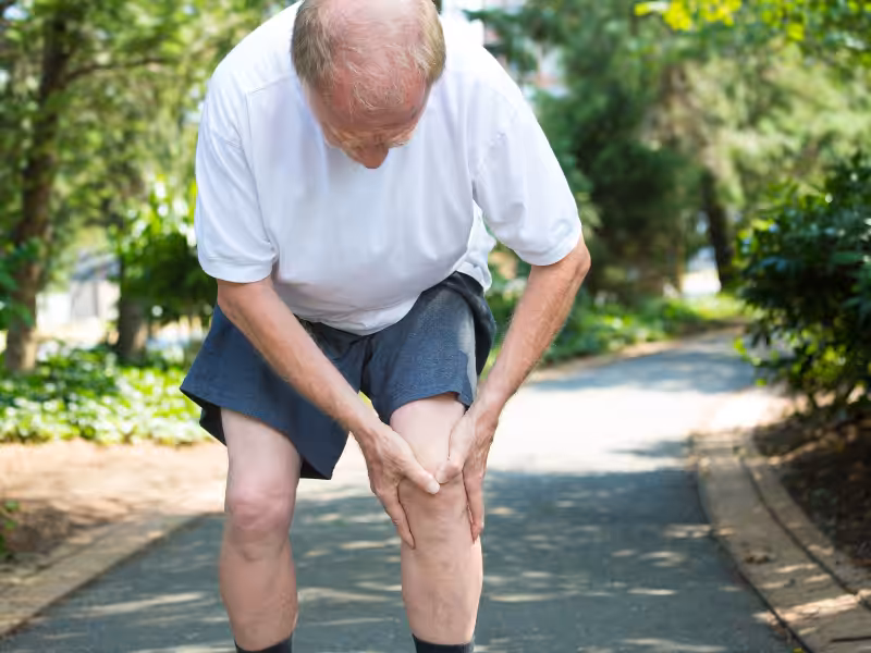 An older man wearing a white shirt and shorts stands on a park path, bending forward and holding his knee in apparent pain, surrounded by greenery.