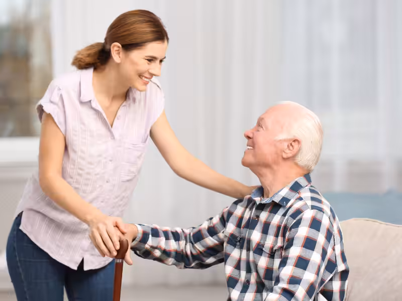 A young woman smiles and talks with an elderly man sitting on a couch, holding a cane. They appear happy and engaged in conversation in a bright, comfortable home setting.