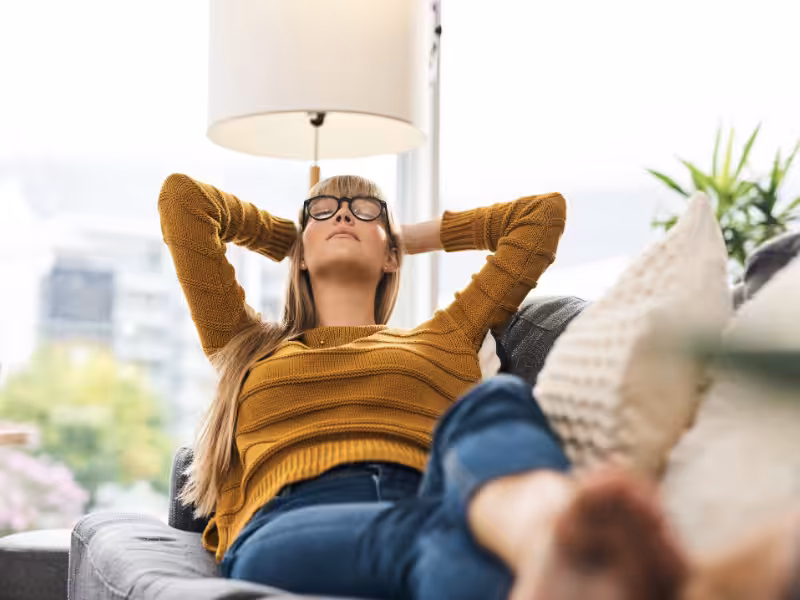 A woman wearing glasses and a mustard sweater relaxes on a gray sofa with her hands behind her head and eyes closed, in a bright living room with large windows and indoor plants.