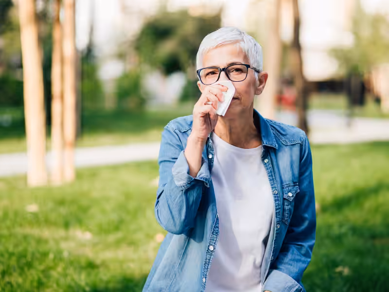 An older woman with short gray hair and glasses sits outside on grass, wearing a denim jacket and white shirt, drinking from a small cup.