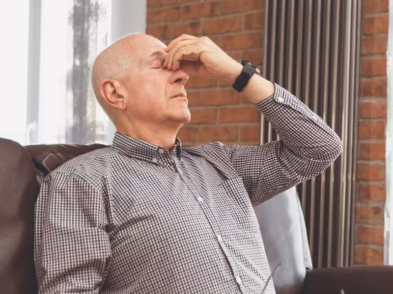 An older man sitting on a couch pinches the bridge of his nose with his eyes closed, appearing stressed or fatigued. He is wearing a checkered shirt and a watch, and is indoors with a brick wall in the background.