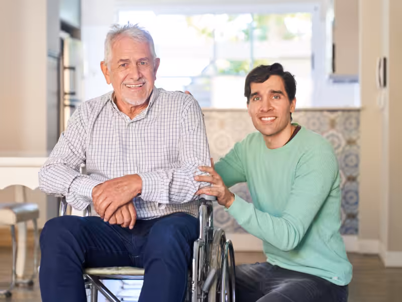 An older man sits in a wheelchair, smiling, while a younger man kneels beside him with a hand on his arm, both looking at the camera in a bright, homey room.