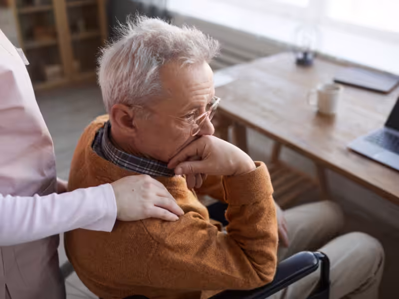 An elderly man sits in a wheelchair, resting his chin on his hand and looking thoughtful. A person beside him gently places a comforting hand on his shoulder. A desk and laptop are visible in the background.