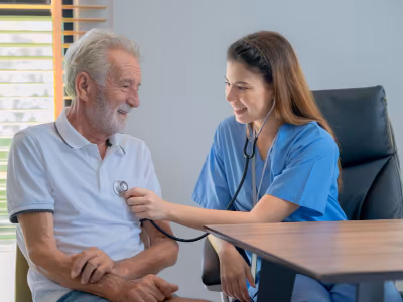 A nurse in blue scrubs uses a stethoscope to check the heartbeat of an elderly man in a white polo shirt. Both are smiling and seated in a bright, comfortable room.