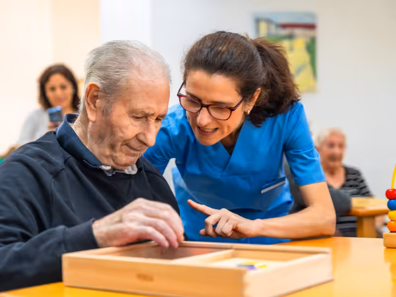 A smiling caregiver in blue scrubs assists an elderly man with a wooden puzzle at a table in a brightly lit room, with other people in the background.