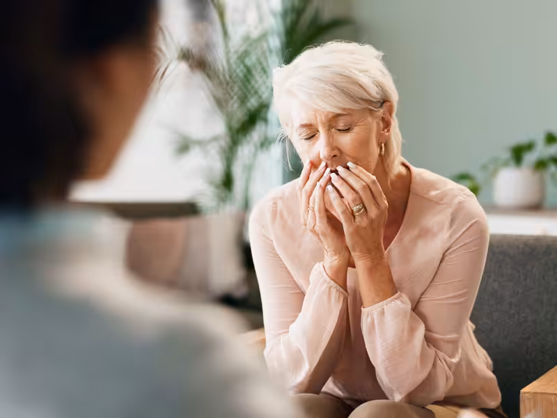 An older woman with short gray hair sits on a chair, holding her hands to her face and appearing distressed or emotional, while another person sits across from her in a softly lit room with plants in the background.