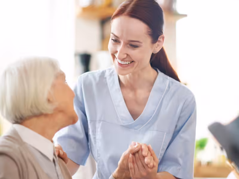 A smiling caregiver in scrubs holds hands with an elderly woman, offering support and comfort in a brightly lit room.