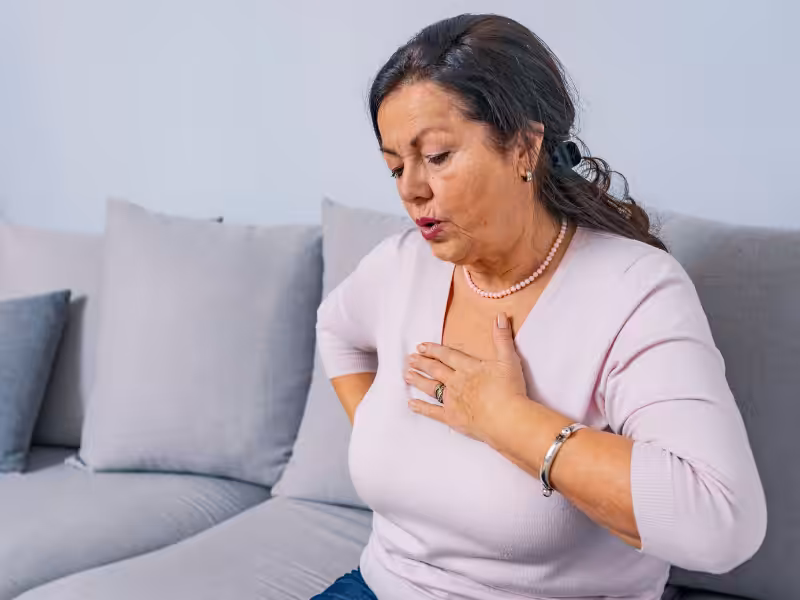 A middle-aged woman sits on a couch, holding her chest with one hand and appearing to have difficulty breathing or experiencing chest pain. She looks concerned and is dressed in a light pink top.