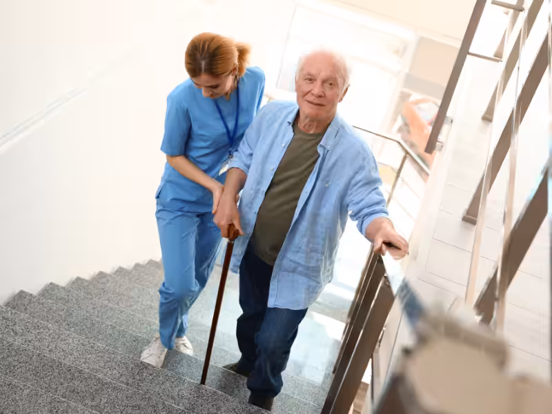 A young female caregiver in blue scrubs assists an elderly man with a cane as he carefully walks up a staircase, holding onto the handrail for support.