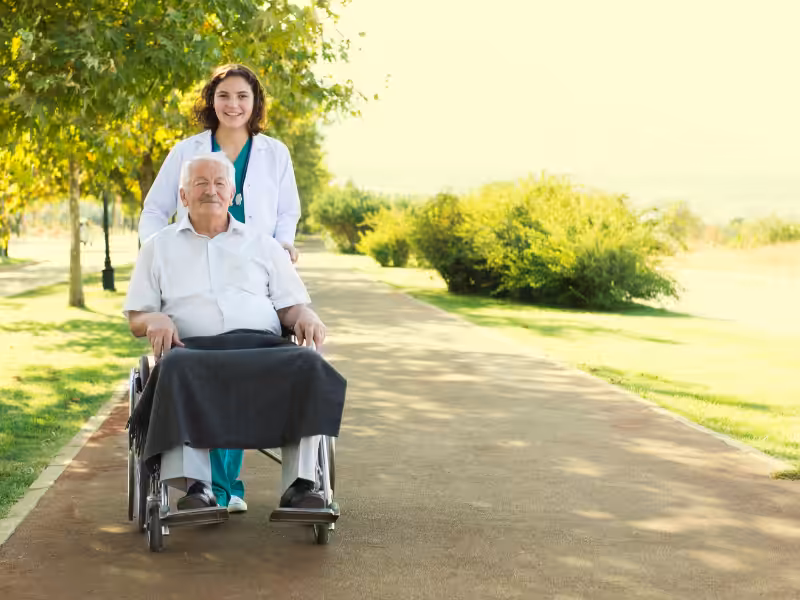 A smiling healthcare worker stands behind an elderly man in a wheelchair on a path in a sunlit park, surrounded by greenery and trees. The man has a blanket on his lap.