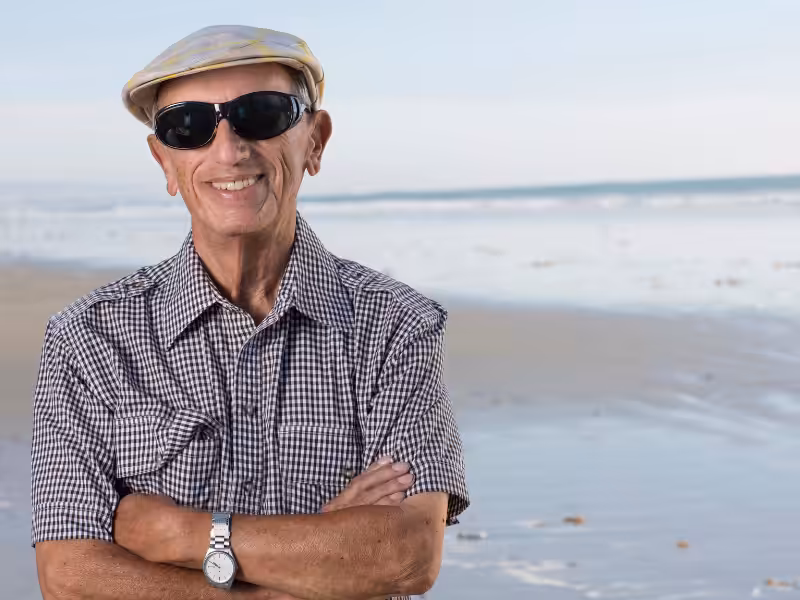 Smiling older man wearing sunglasses, a checked shirt, a wristwatch, and a cap stands with arms crossed on a sandy beach with gentle waves and blue sky in the background.