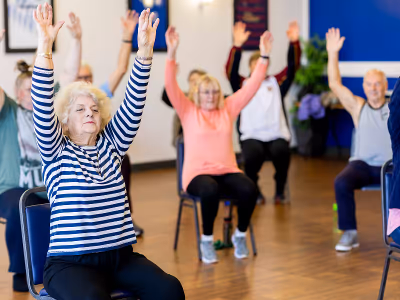A group of older adults is seated in chairs, participating in an exercise class with their arms raised overhead in a bright indoor room.