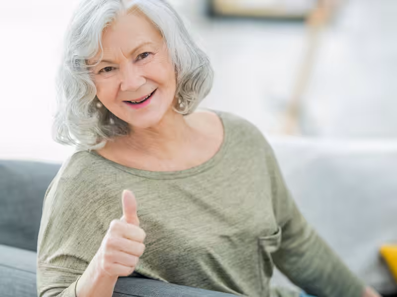 Smiling older woman with gray hair and a green top sits on a couch and gives a thumbs up gesture, expressing positivity and approval.