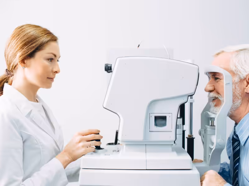 An eye doctor conducts an eye exam for an older man using a modern ophthalmic device in a clinical setting. The man is sitting and facing the machine, while the doctor adjusts the controls.