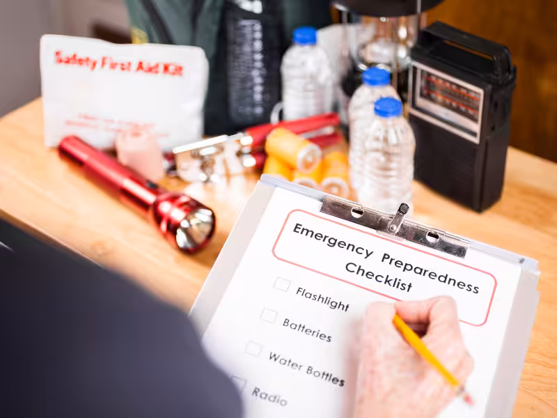 A person fills out an emergency preparedness checklist on a clipboard. Nearby are a red flashlight, batteries, bandages, bottled water, a first aid kit, and a radio, all on a wooden table.