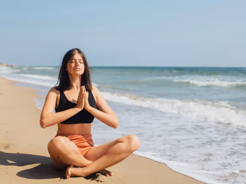 A woman sits cross-legged on a sandy beach near the shoreline, eyes closed and hands pressed together in a prayer pose, meditating under a clear sky with gentle waves in the background.