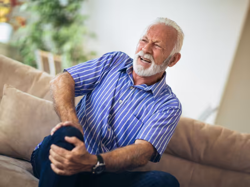 An elderly man with a white beard sits on a couch, wearing a blue striped shirt, and holds his knee with both hands while grimacing in pain.