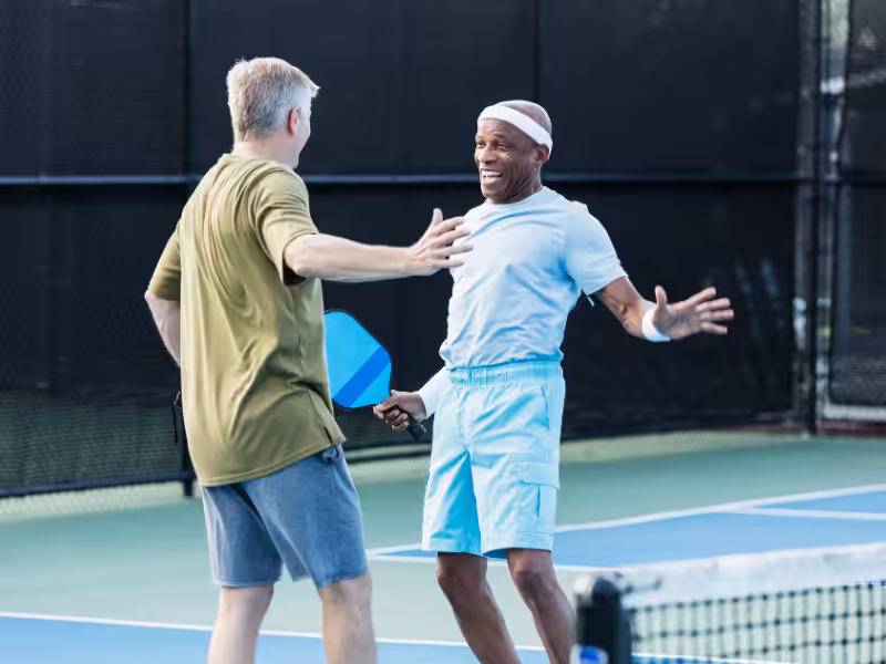 Two men on a pickleball court, smiling and reaching out for a celebratory high five, one holding a paddle. Both are wearing casual athletic clothing and appear happy and energetic.