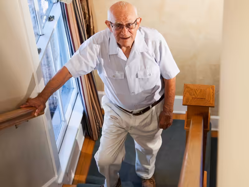 An elderly man wearing glasses, a white short-sleeve shirt, and white pants is walking up a staircase, holding the handrail and looking up, with sunlight coming through a window beside him.