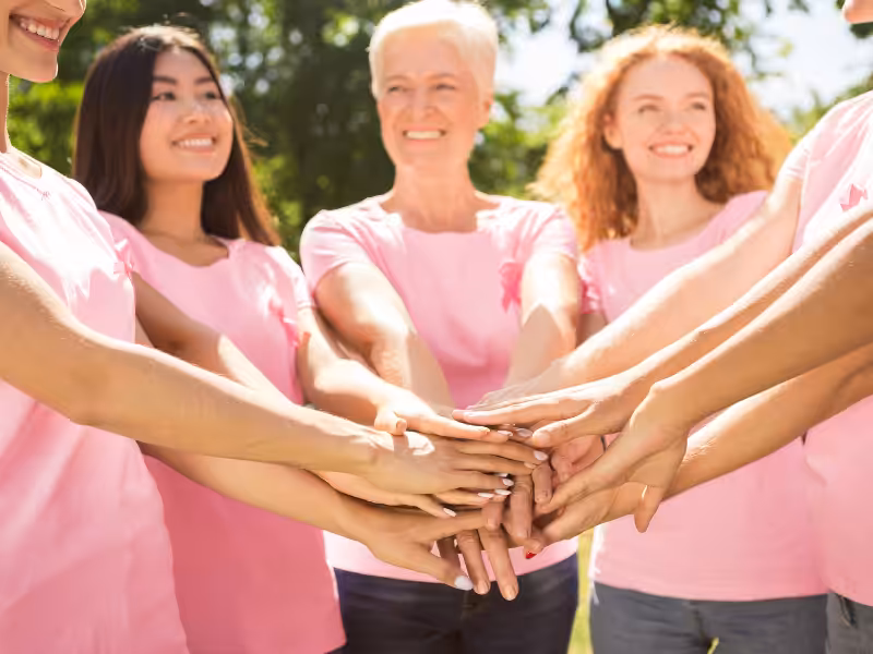 A group of women wearing pink t-shirts stand in a circle outdoors, smiling and stacking their hands together in the center, showing unity and support.