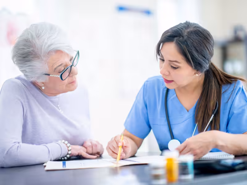 A nurse in blue scrubs discusses paperwork with an older woman who is wearing glasses and a light purple sweater. They sit at a table with medical documents and pills, looking focused on the discussion.