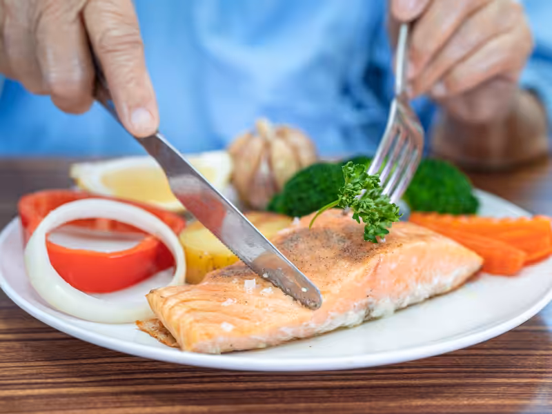 A person uses a knife and fork to cut a cooked salmon fillet served with parsley, broccoli, carrots, tomato, onion, and a lemon wedge on a white plate.
