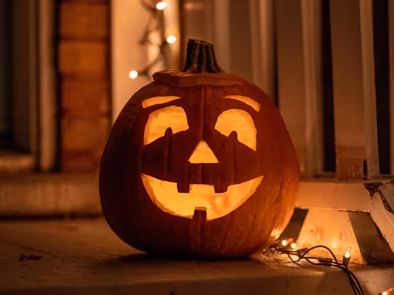 A carved pumpkin with a glowing, smiling face sits on a porch, illuminated by string lights, creating a festive Halloween atmosphere at night.