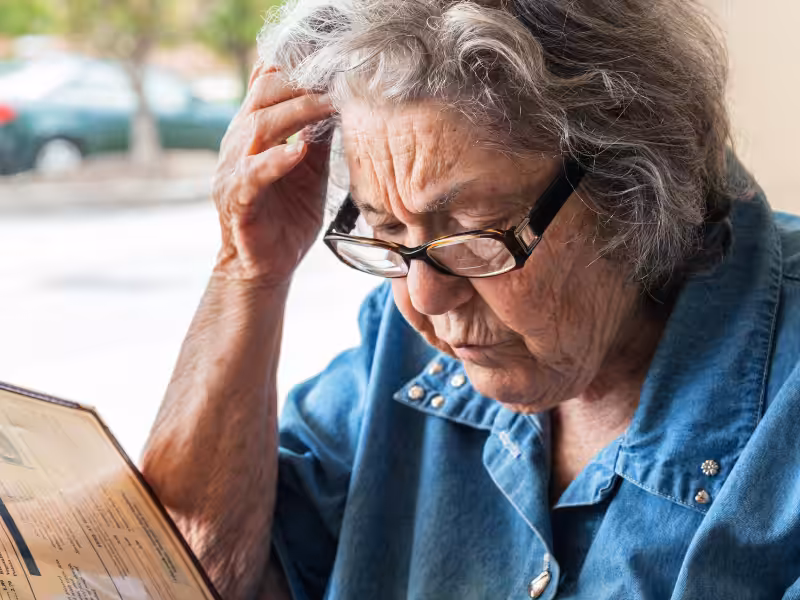 An elderly woman wearing glasses and a blue denim shirt intently reads a menu, holding her head in concentration. She is seated indoors near a window with blurred outdoor scenery in the background.