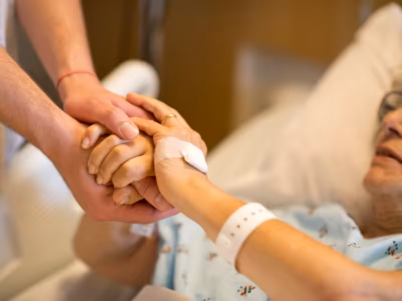 A patient in a hospital bed holds hands with another person. The patient wears a hospital gown and ID bracelet and has an IV in their hand, while the other person offers comfort and support.