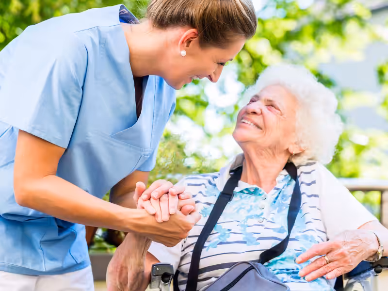 A smiling healthcare worker in blue scrubs holds hands with an elderly woman in a wheelchair outdoors, both looking at each other and smiling warmly. Lush greenery is visible in the background.
