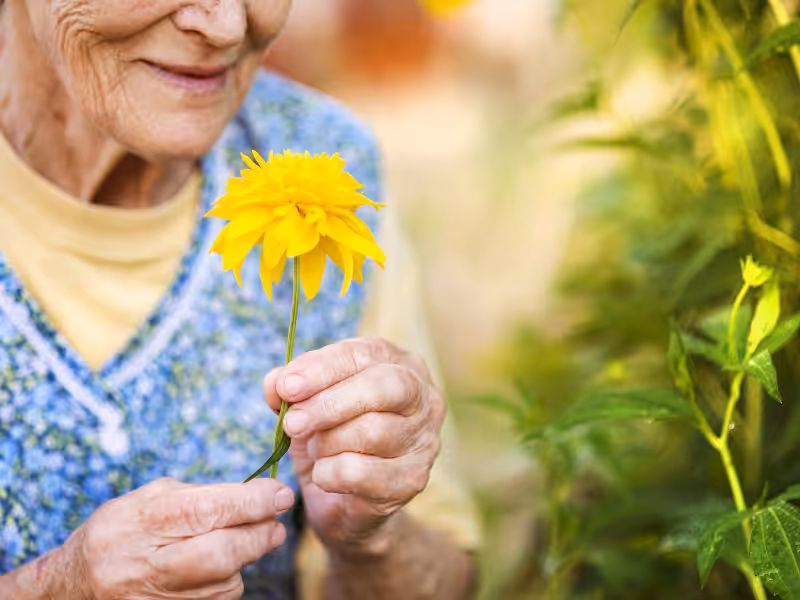 An elderly woman, wearing a floral-patterned dress, gently holds and admires a bright yellow flower while smiling in a garden setting.