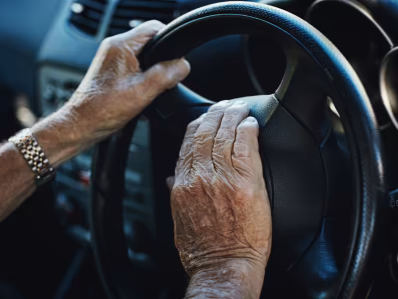 Close-up of an elderly persons hands on a car steering wheel, one hand pressing the horn. The dashboard and part of the cars interior are visible in the background.