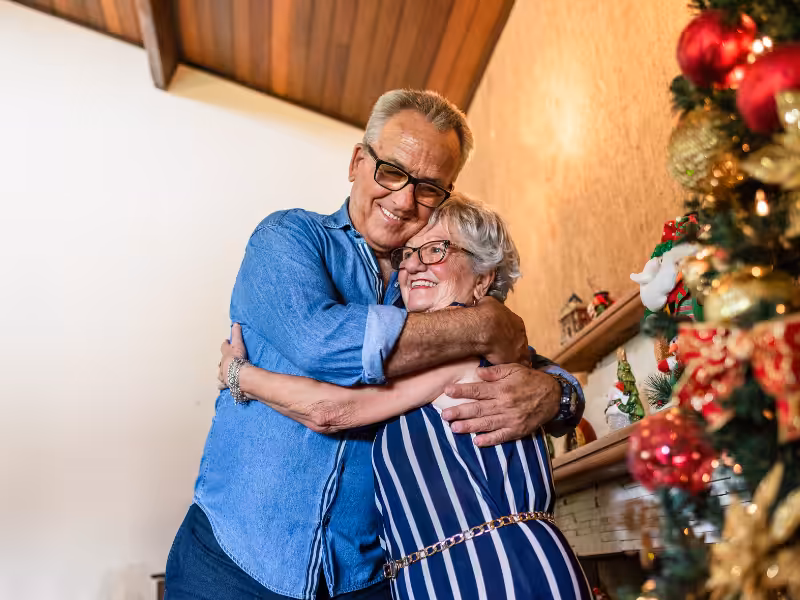 An elderly couple hugs and smiles beside a decorated Christmas tree in a cozy room with a wooden ceiling, creating a warm and festive atmosphere.