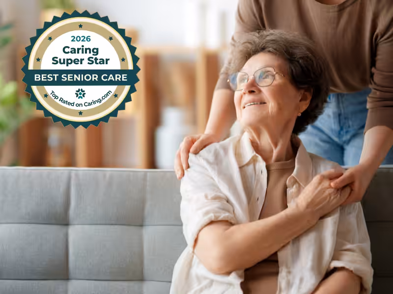 An elderly woman sits on a couch, smiling and holding hands with a younger person standing beside her. A badge reads 2026 Caring Super Star: Best Senior Care, Top Rated on Caring.com.