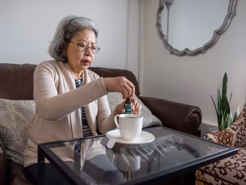 An older woman sitting on a couch adds drops from a small bottle labeled CBD Oil into a cup on a saucer placed on a glass coffee table in a cozy living room.