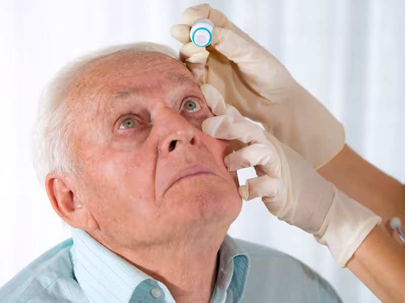 An older man tilts his head back while a gloved hand holds his eyelid and administers eye drops. The background is softly blurred, emphasizing the medical procedure.