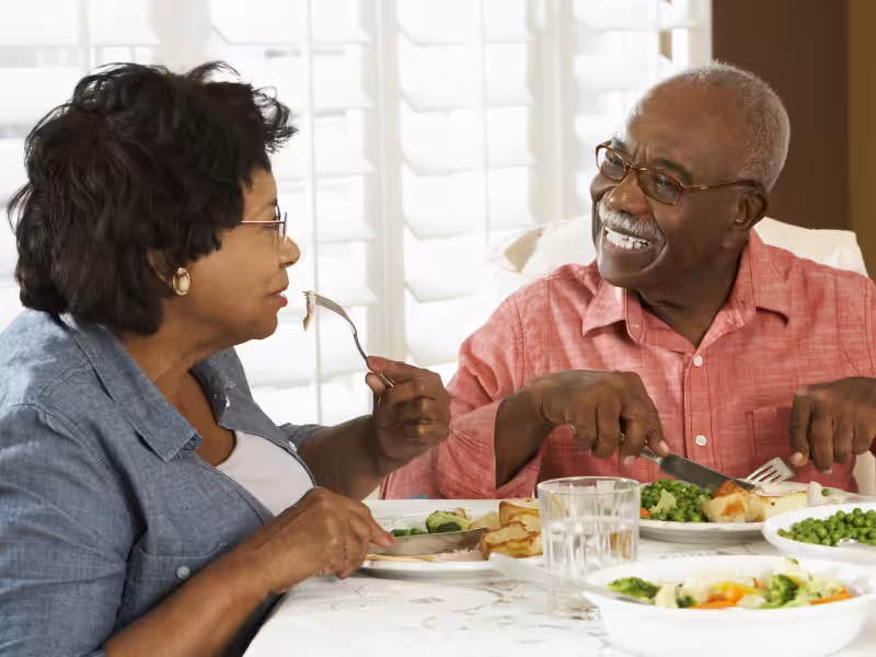 An elderly couple sits at a dining table, smiling and talking while enjoying a meal together. Plates of food and glasses of water are in front of them, and sunlight comes through window blinds in the background.
