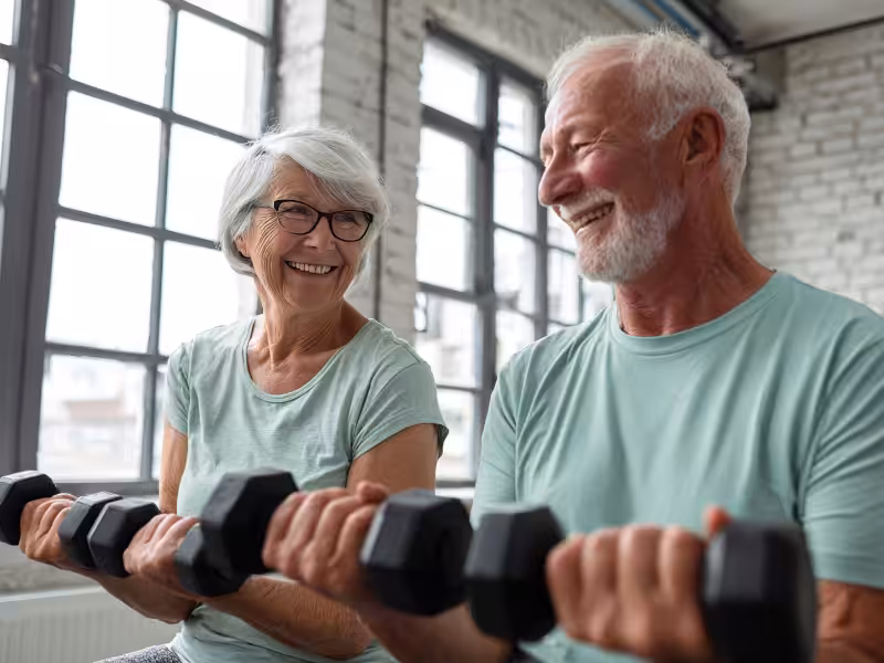 Two smiling older adults in light blue shirts sit indoors by large windows, exercising with dumbbells. They look at each other happily, suggesting they are enjoying their workout together.